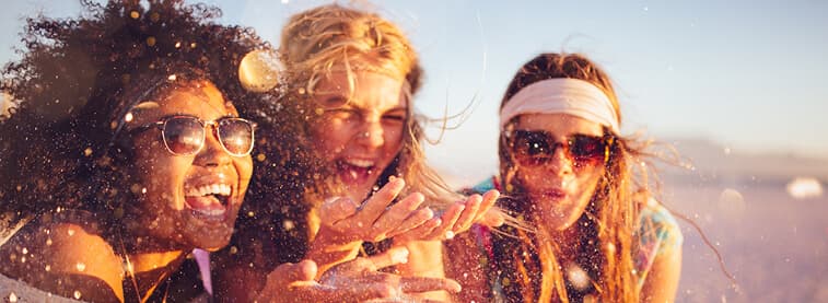 Three young women, left and right both wearing sunglasses, all laughing in the sunshine