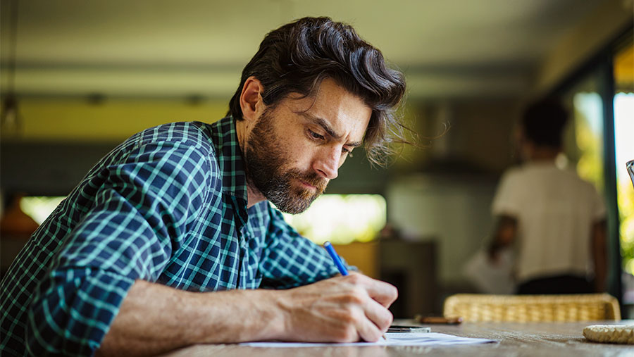 Man in a blue check shirt sat at a table looking down at the form he is filling out