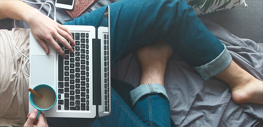 Male with laptop on his lap with cup of tea in his right hand and left hand hover keyboard