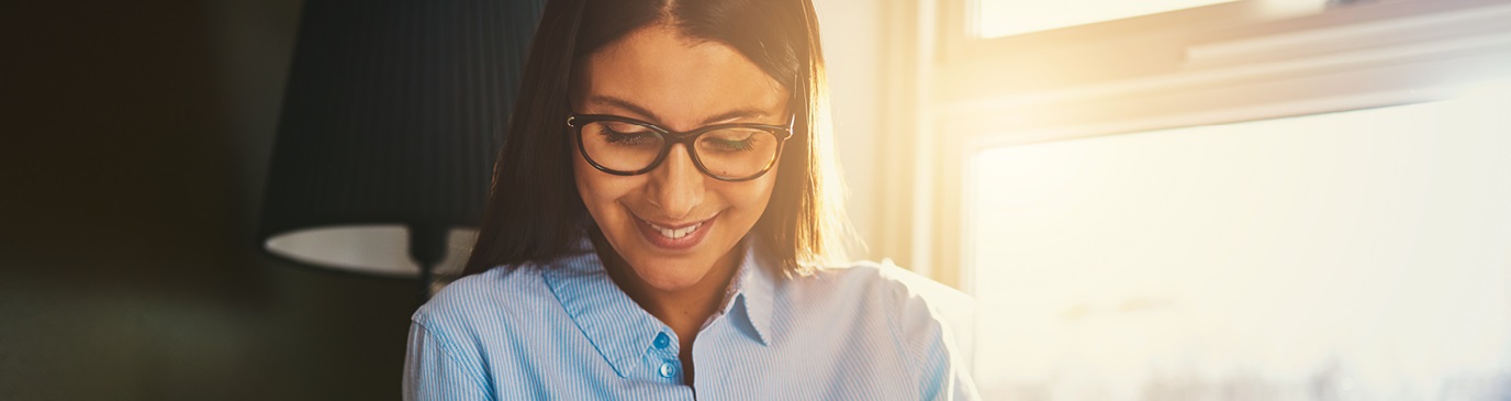 Young smiling woman in light blue shirt wearing glasses, sat in a sunlit lounge, looking at laptop