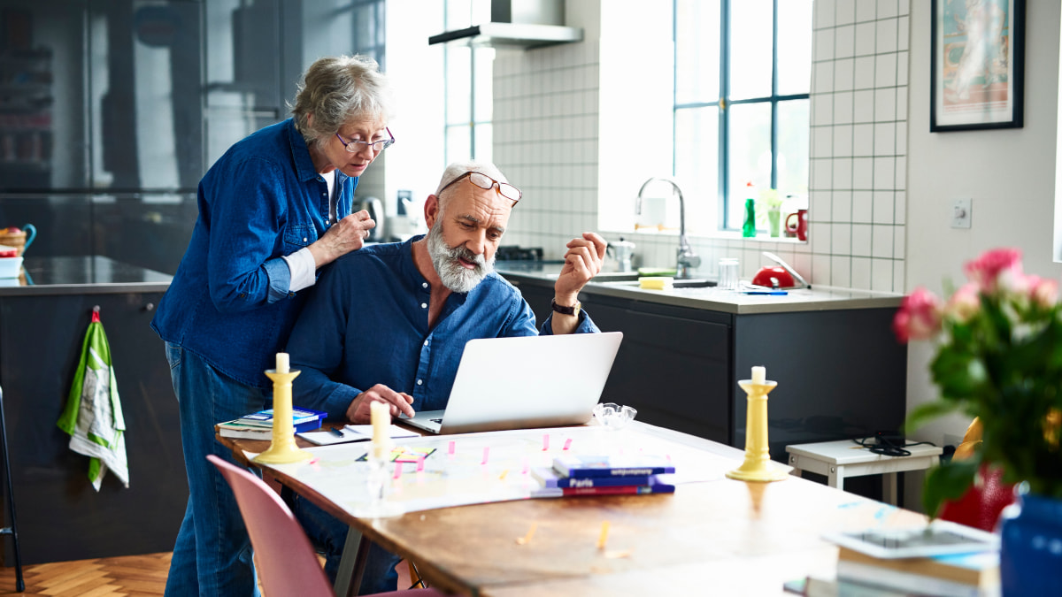 Couple in the kitchen, man sat at table at a laptop and female behind him both looking at the laptop