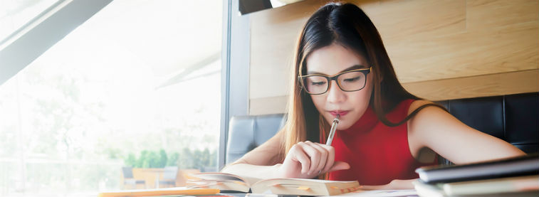 Woman sat in a well-lit café, holding pen to her mouth while reading from a book for her studies