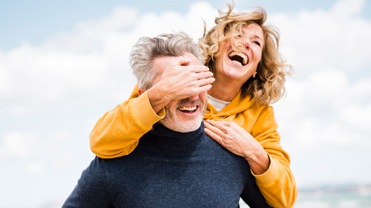 Lady behind bearded male with her right hand over his eyes and left hand on his left shoulder