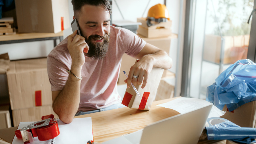 Bearded male smiling talking on his mobile sat at a table with a parcel under his arm